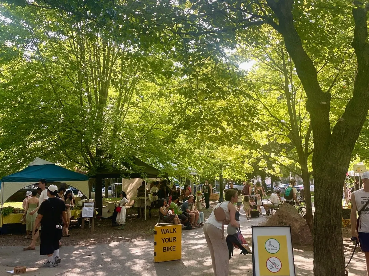 farmers' market in a park under the canopy of green trees. People walking and shopping at the various booth.