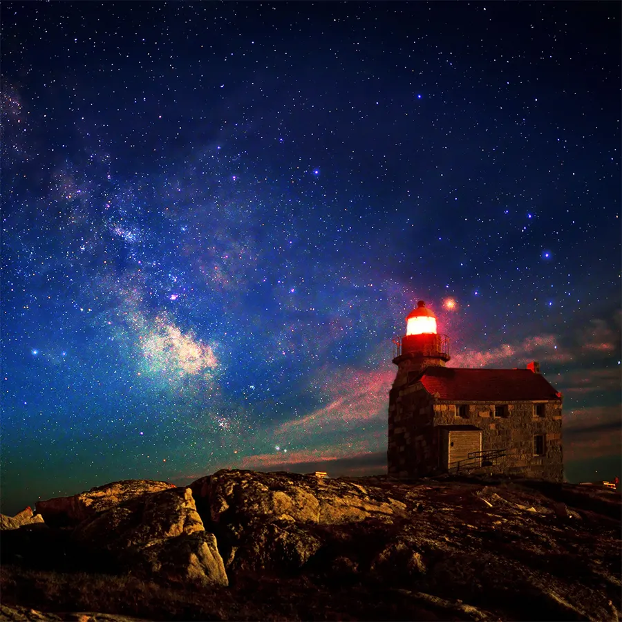 Stone lighthouse sat upon rocks with northern lights across the night sky