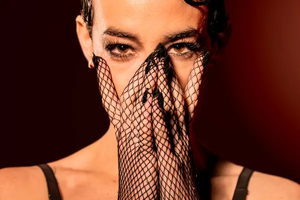 A close-up portrait of a performer covering the lower half of their face with hands wearing black fishnet gloves.