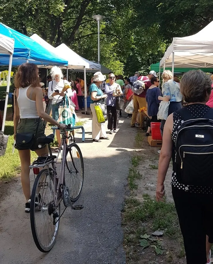 Busy path in a park with vendor tents. Customer walking with a bike. Other customers shopping.