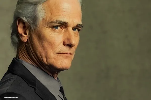 A side-profile portrait of a grey-haired man in a dark suit looking intently at the camera against a textured backdrop.