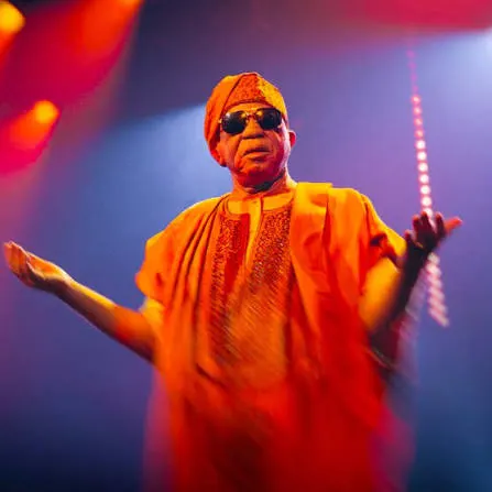 A performer in a bright orange outfit and sunglasses gestures with raised arms on a colourful, illuminated stage.