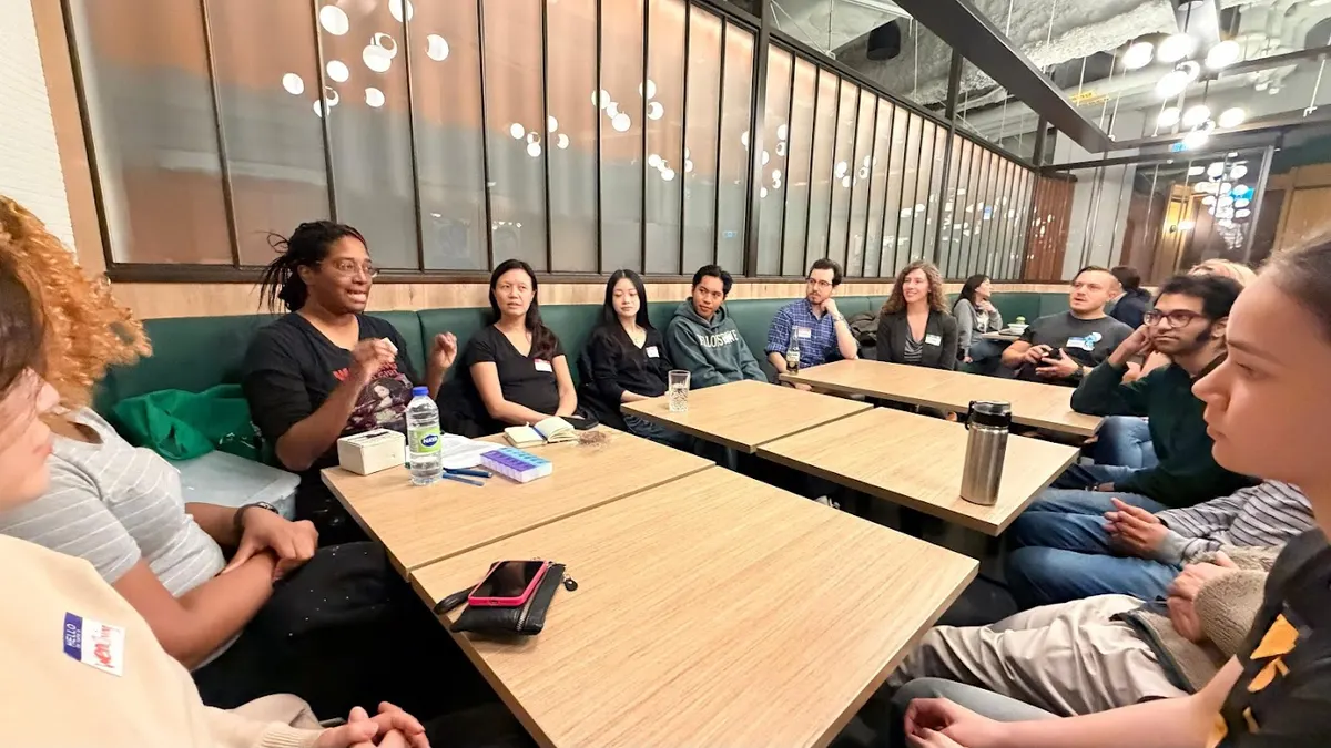 A group of 12 people are seated around square tables, listening to someone give instructions near the left