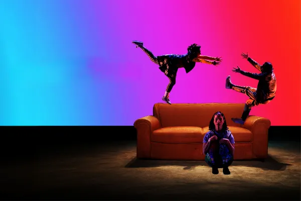 Two children play on top of an orange couch. One begins to fall. Another child sits at the base of the couch.