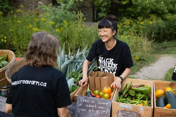 two staff at the booth with fresh seasonal produce in boxes and baskets. Asian woman smiling. Other facing back.