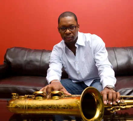 Ravi Coltrane sits on a sofa. He wears a white shirt and his saxophone is placed in front of him on the table.