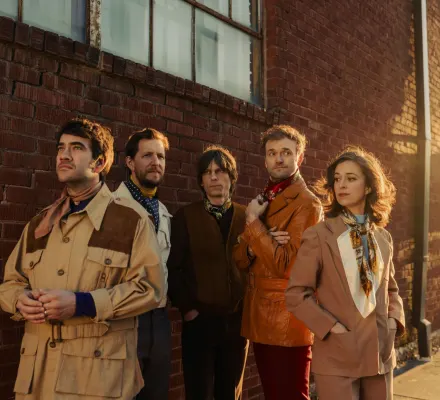 A group of people dressed in earth tones stand next to a red brick wall.