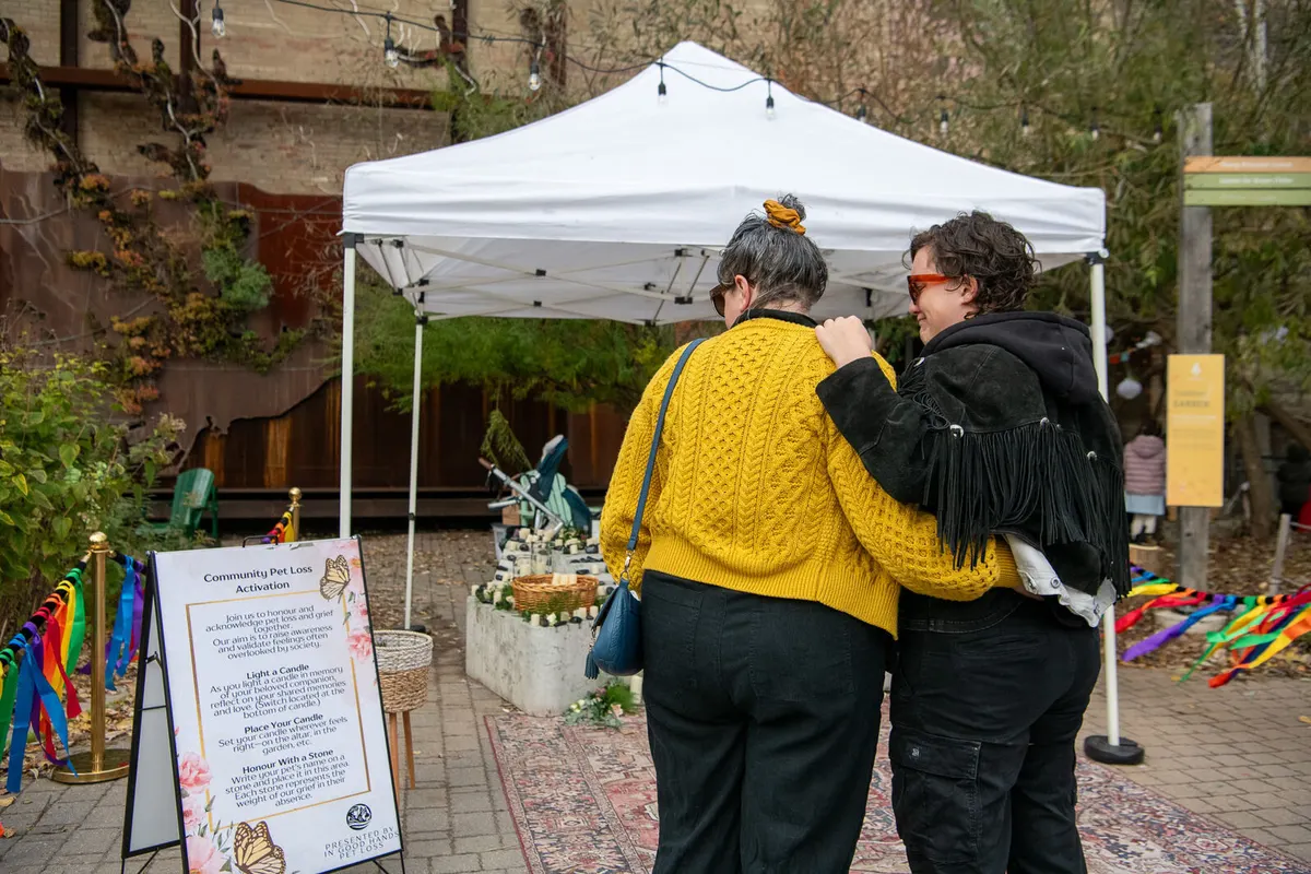 Outdoors, under a white tent, a pet memorial is watched by a woman in a yellow sweater and a woman in black, hugging