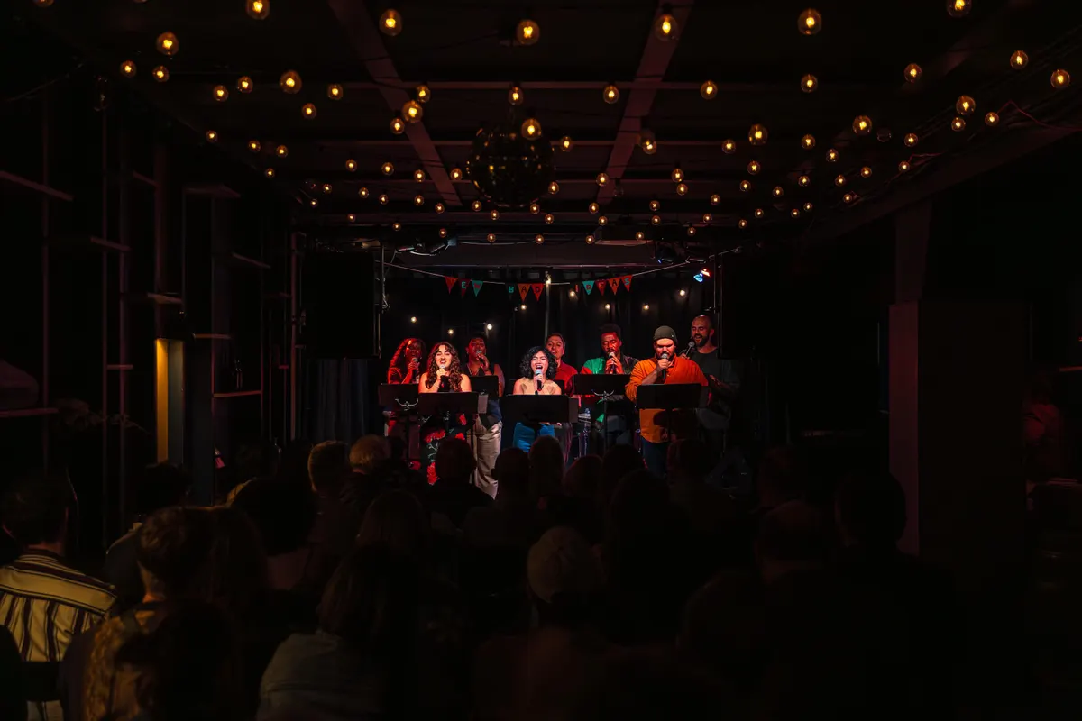 Eight performers stand on a well-lit stage with microphones in hand.
