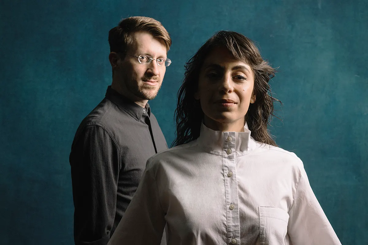 One female musician in white top and one male musician with glasses in a black top, on a blue background