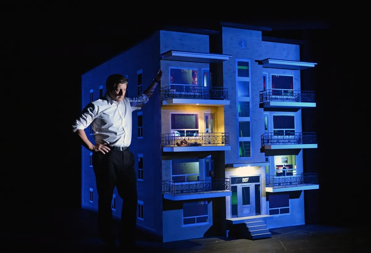 Robert Lepage on stage, standing next to a scale model of a 4-floor apartment complex with an 887 on the front door.